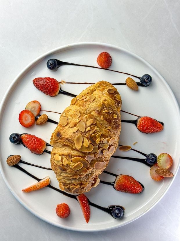 An almond croissant coated in flaked almonds, ringed with chocolate sauce, berries and whole almonds