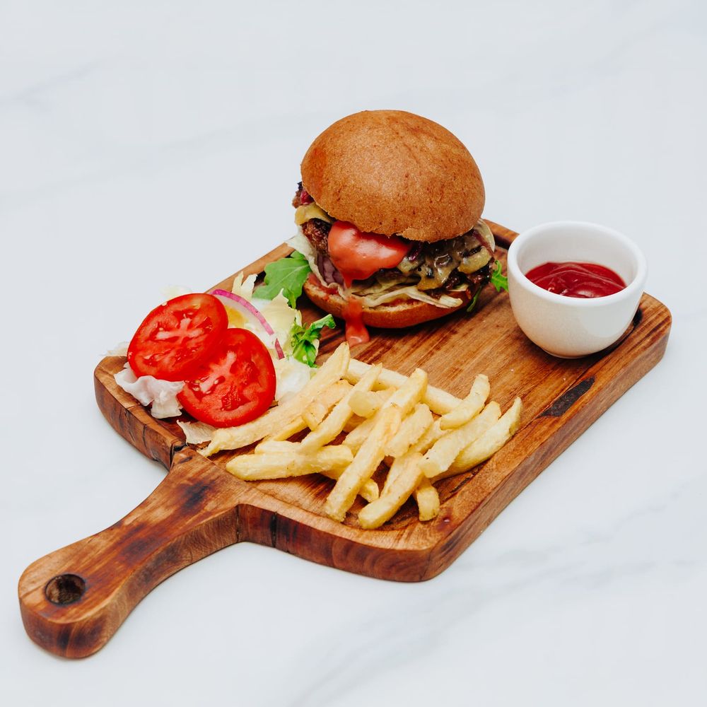 A cheeseburger on a wooden board with fries, sliced tomato, salad and a ramekin of ketchup