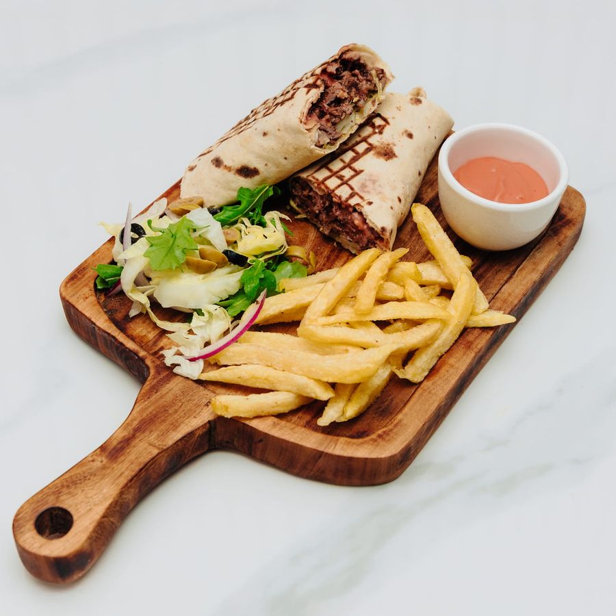 A grill-marked shawarma wrap cut in half on a wooden board with fries, salad and a ramekin of sauce