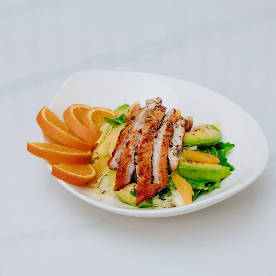 Sliced grilled chicken fanned over salad greens with avocado and orange segments in a white bowl