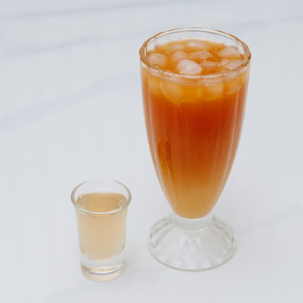 A tall footed glass of iced amber juice with a small shot glass alongside
