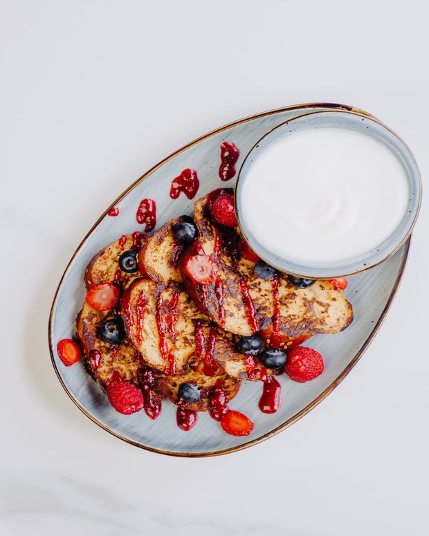 Slices of griddled French toast on an oval platter with berry coulis, fresh berries and a bowl of cream