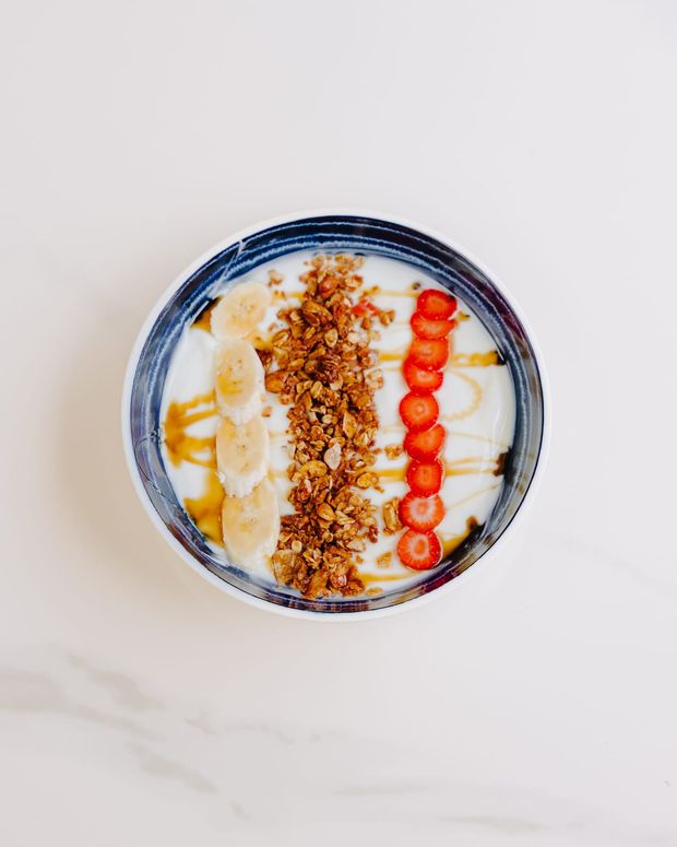 A bowl of thick yoghurt striped with banana, granola and sliced strawberries, drizzled with honey