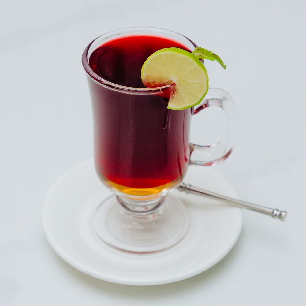 A footed glass mug of deep red hibiscus tea with a lime wheel and mint on the rim
