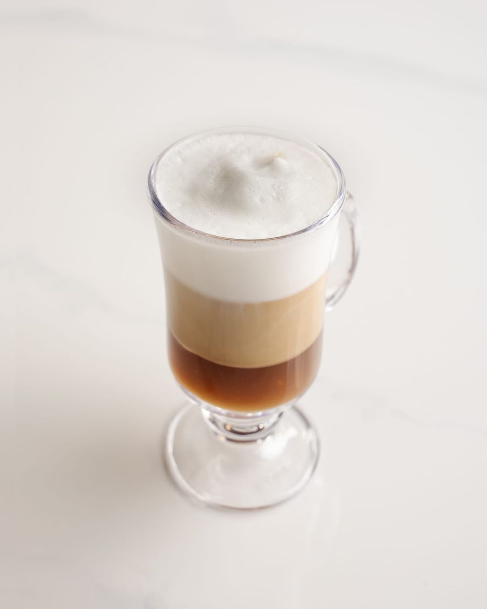 A layered macchiato in a clear footed glass showing espresso, milk and a thick foam cap