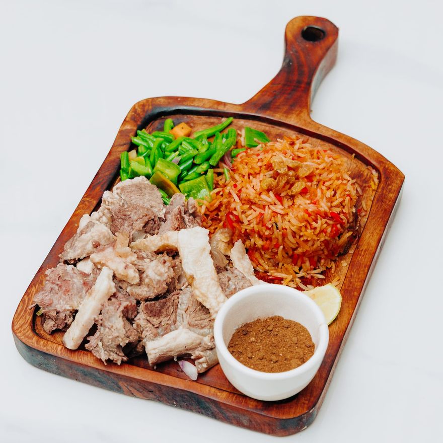 Slow-cooked meat on a wooden board with spiced rice, sautéed vegetables, a lemon wedge and a dish of spice