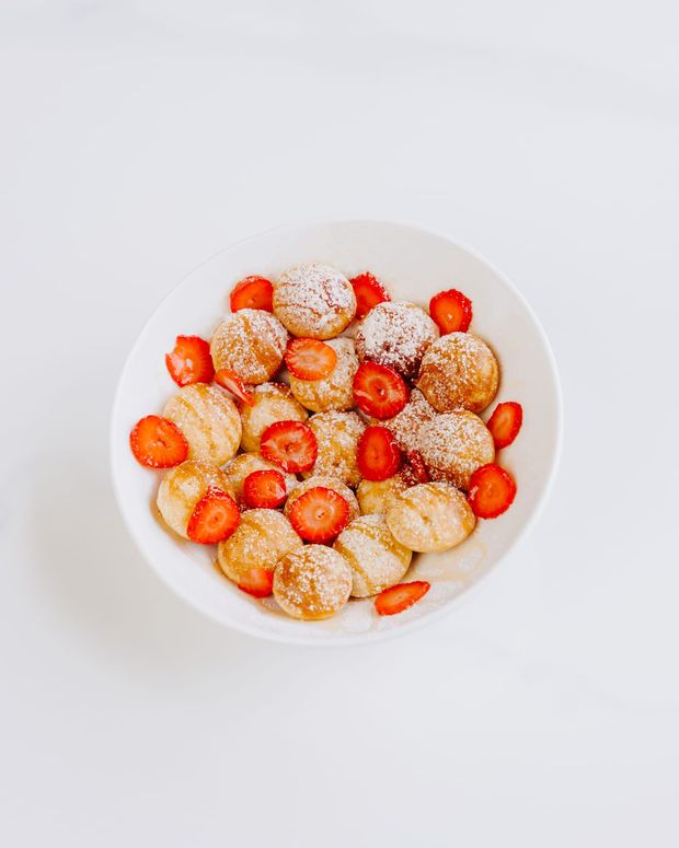 A bowl of golden mini pancake bites dusted with icing sugar and ringed with halved strawberries