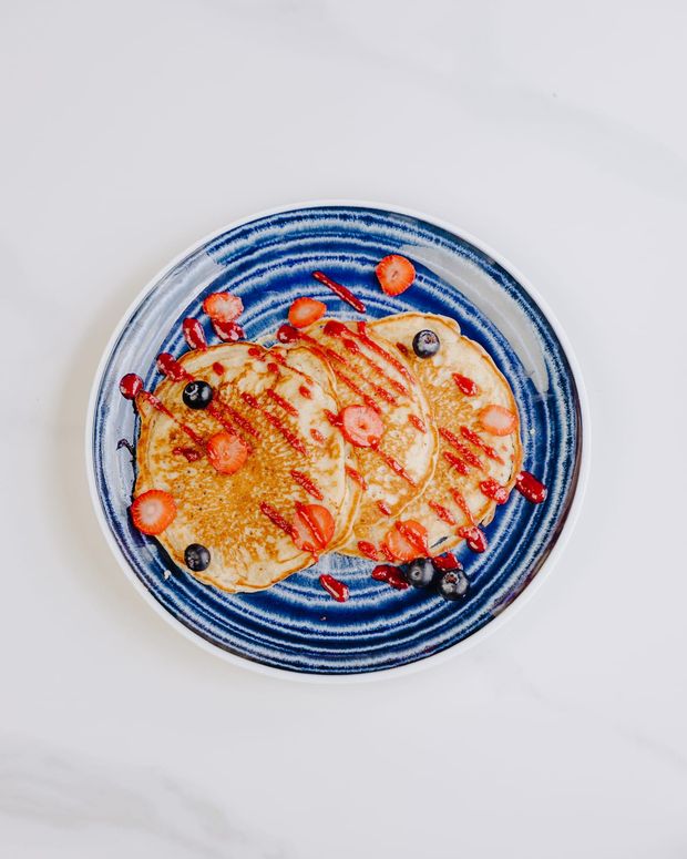 Three pancakes fanned across a blue stoneware plate with strawberry coulis, sliced strawberries and blueberries