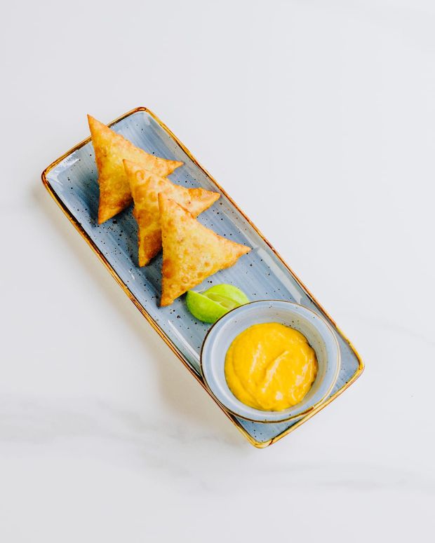 Three golden samosas fanned across a blue platter with lime wedges and a ramekin of mango dipping sauce