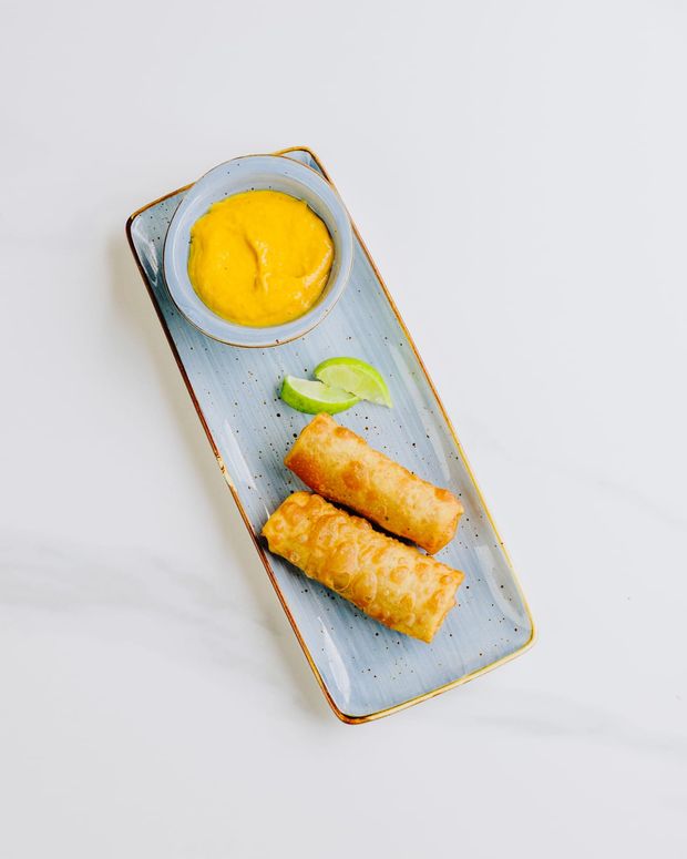 Two crisp golden spring rolls on a blue platter with lime wedges and a ramekin of mango dipping sauce