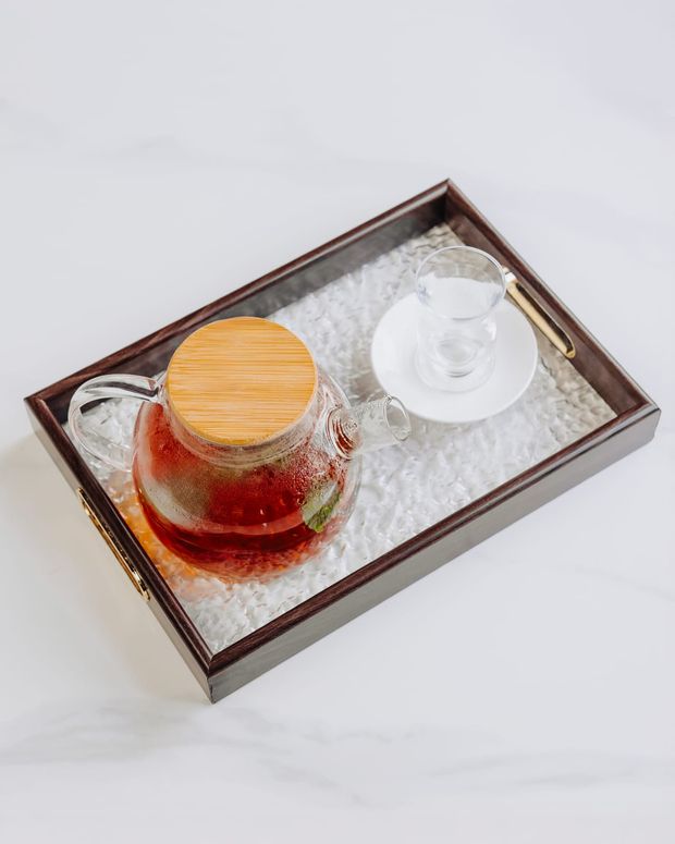 A glass teapot of amber mint tea on a wooden tray with an empty tulip glass and gold spoon
