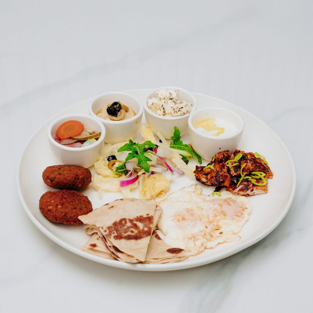A large breakfast platter with a fried egg, patties, chapati, salad and small dishes of hummus, feta and yoghurt
